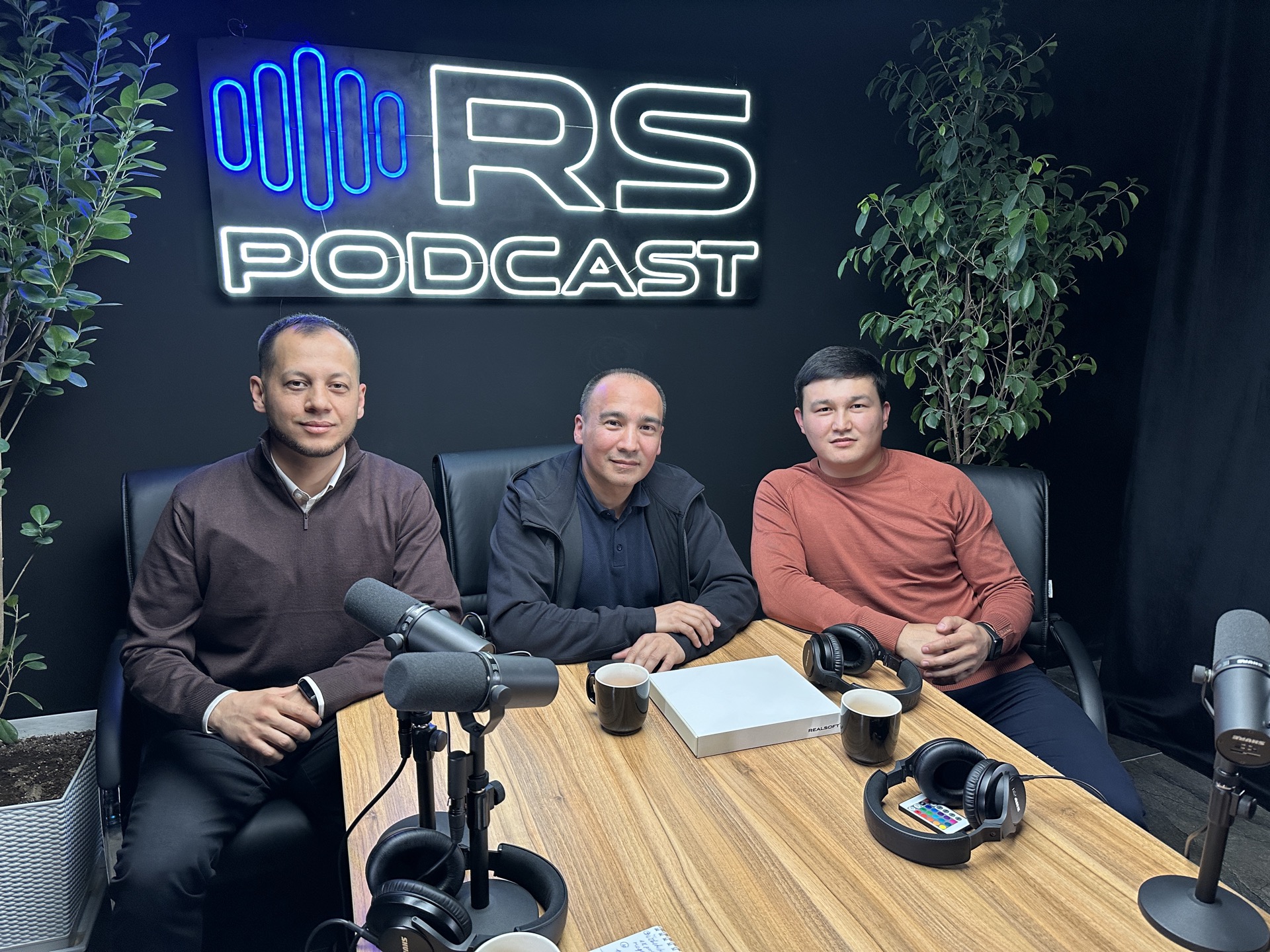 Khurshid Normurodov hosting the RS Podcast with two guests at the studio table, RS Podcast neon sign behind them